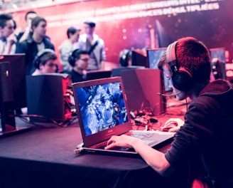 Several people are seated and focused on computer screens, wearing headsets, in an environment that appears to be a gaming tournament or esports event. The room is dimly lit with neon lighting, highlighting screens and creating an intense atmosphere.