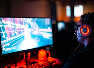 A person wearing headphones is sitting in front of a computer monitor. The screen displays a vibrant, illuminated video game scene, while the person appears focused on gaming. The ambient lighting is dark with red and blue tones, and a backlight from the monitor highlights the headphones.