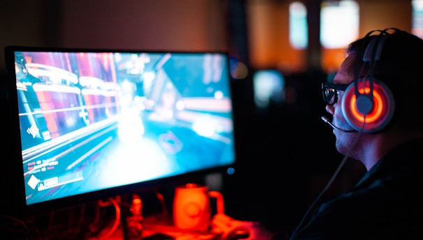 A person wearing headphones is sitting in front of a computer monitor. The screen displays a vibrant, illuminated video game scene, while the person appears focused on gaming. The ambient lighting is dark with red and blue tones, and a backlight from the monitor highlights the headphones.