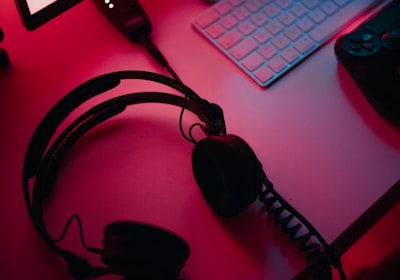 A dimly lit gaming setup with a monitor displaying a game's menu on the screen. A mechanical keyboard sits in front of the monitor. A pair of over-ear headphones and a game controller rest on the desk. The scene is bathed in a red and blue light, creating a moody gaming atmosphere.