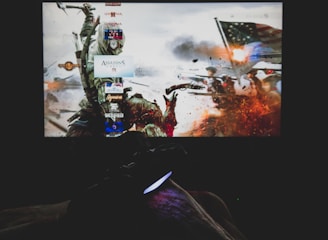 A dimly lit room with a television screen displaying a dynamic, action-packed video game interface featuring a historical battle scene. The foreground shows the silhouette of a gaming console controller held by a person.