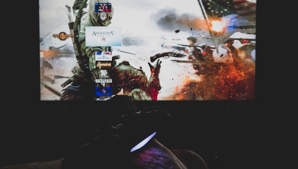 A dimly lit room with a television screen displaying a dynamic, action-packed video game interface featuring a historical battle scene. The foreground shows the silhouette of a gaming console controller held by a person.