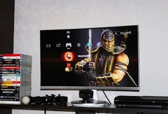 A flat-screen TV displays a video game interface featuring a character from Mortal Kombat in the center. Beside the TV is a stack of video game cases for the PlayStation 3, including titles like FIFA 14 and Grand Theft Auto. Below the TV, there is a gaming console, controllers, and other gaming equipment.