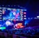 A large esports arena featuring a massive screen displaying a video game, surrounded by sponsor logos and team names. The auditorium is filled with spectators watching the gaming event. Neon blue and purple lights illuminate the stage, creating a vibrant atmosphere.