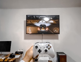 A person is holding a gaming controller in front of a flat-screen television displaying a racing video game. The room has a computer setup on a wooden desk with cables, a desktop computer monitor, and other accessories. The focus is on the interaction between the person and the gaming environment.
