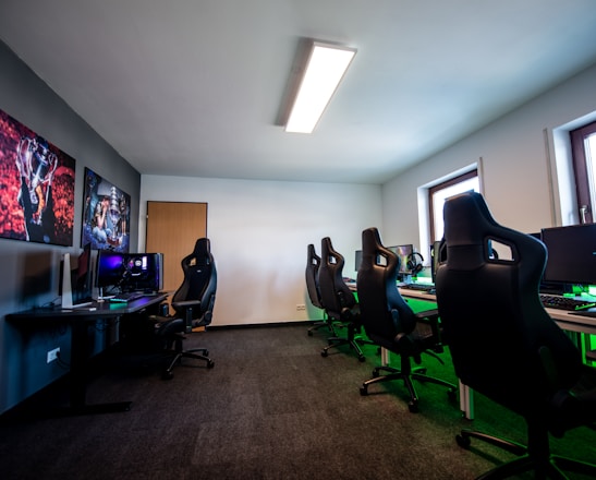 A well-lit gaming room features multiple gaming stations with high-backed chairs and monitors. The room has a minimalist setup with green and black color tones. Large posters adorn the walls, and the room appears clean and organized.