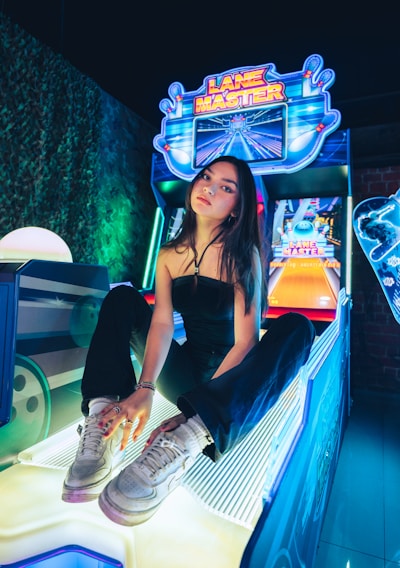 A person sitting on an arcade game machine platform with neon lights in the background. The arcade game is called 'Lane Master' and features a colorful design with bowling-themed imagery.