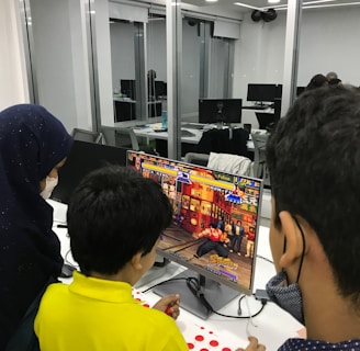 A group of children is gathered around a desktop computer, engaged in playing a colorful, action-packed arcade game displayed on the monitor. The setting appears to be a modern, well-lit office or gaming space with large glass windows, showing multiple computer workstations in the background.