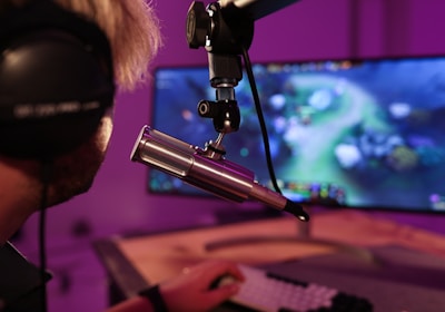 A person wearing headphones is seated in front of a computer screen, engaged in a video game. A microphone is positioned prominently in front of them, suggesting a streaming or recording setup. The background is illuminated with soft purple lighting, creating a cozy and immersive atmosphere.