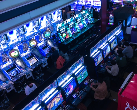 A bustling arcade filled with rows of gaming machines, each with vibrant displays and controls. Several people are seated at these machines, deeply engaged in playing games. Bright fluorescent lights cast a blue and neon glow across the room, enhancing the lively and energetic atmosphere.