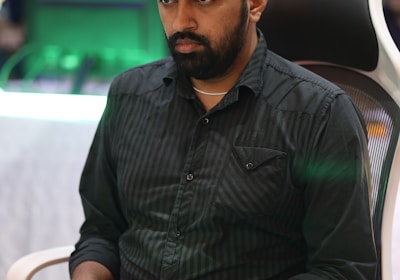 A person is seated in a chair, focused intently on something, with hands positioned on a gaming console or arcade controller. The setup appears to be indoors, with visible screens and lighting in the background, suggesting an electronic gaming environment.